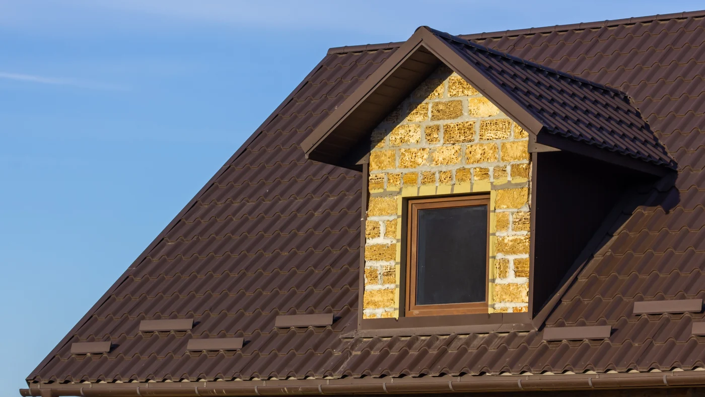 Close-up of a home with a brown metal roof with a small gable, brick siding, and a window on it