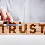 A man’s hand places the final wooden block, building a small wall, and the word “trust” can be seen on the front side