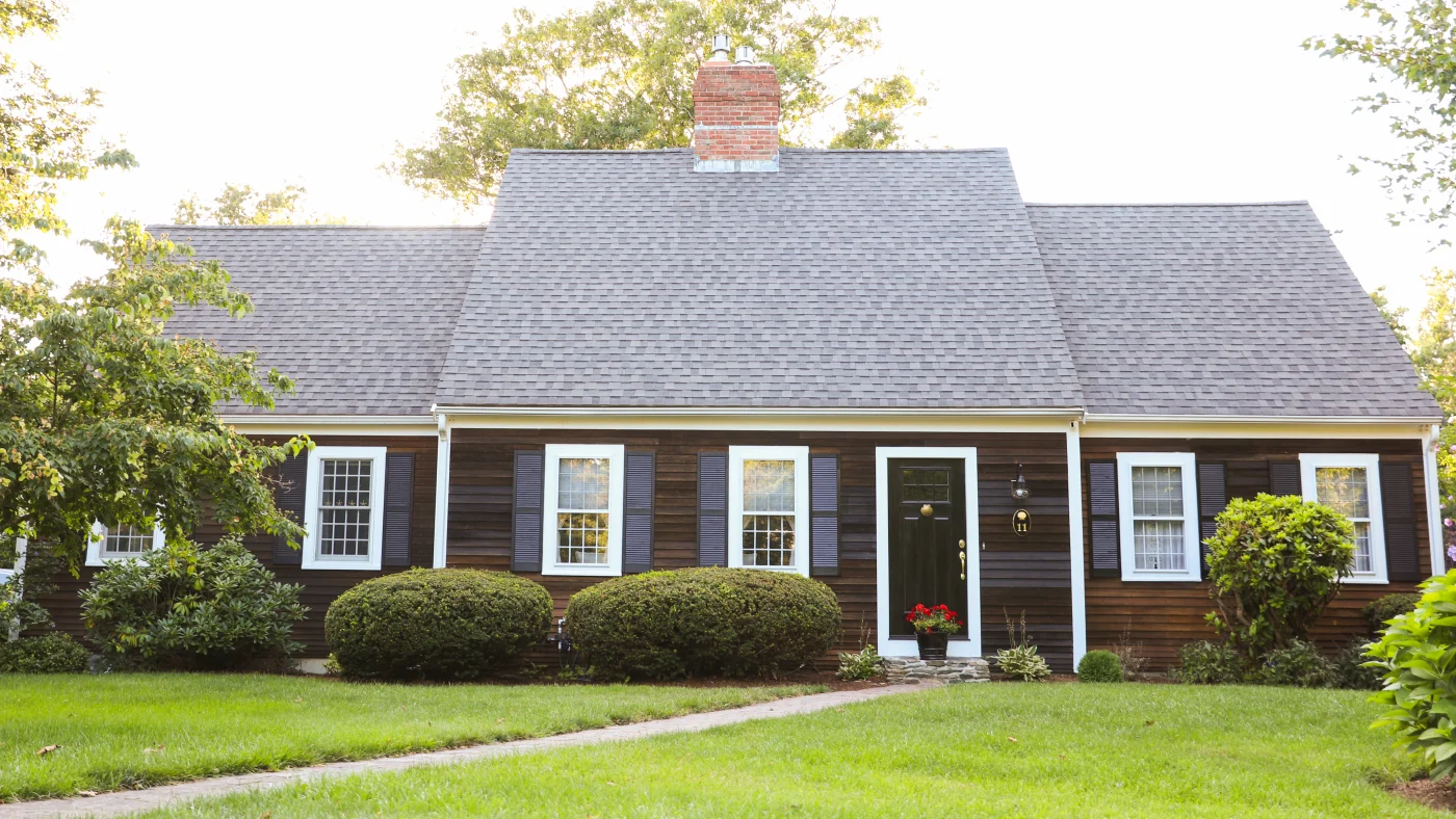 Exterior view of a home with a steep-sloped dark asphalt shingle roof, brown horizontal siding, and white windows with dark shutters on them.