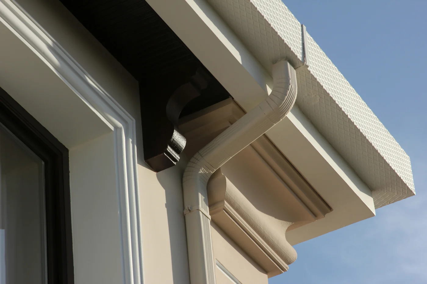 A close-up view of a light-colored gutter with a gutter guard and downspout on a building with light siding and dark trim against a blue sky.