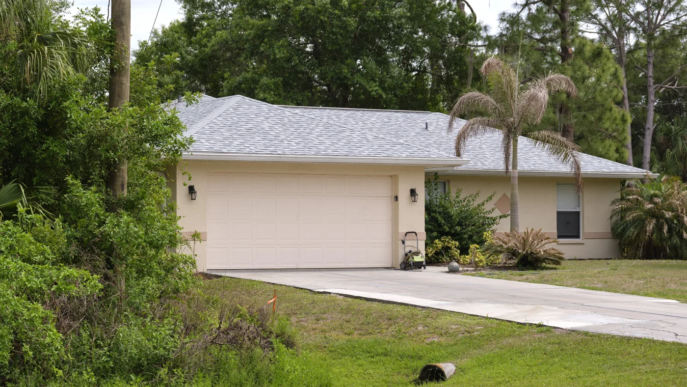 Wide Garage Double Door And Concrete Driveway Of New Modern American House. A single-story house with a light-colored exterior and a gray shingled roof, featuring a two-car garage with a beige door.