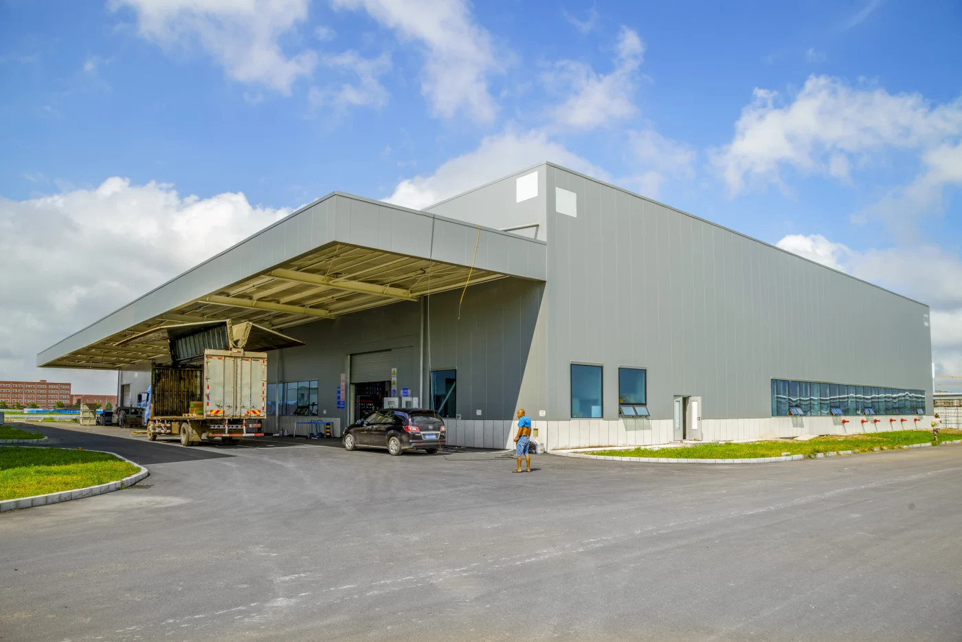 A wide exterior view of a large, modern, grey industrial or warehouse building under a blue sky with some clouds.