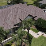 Aerial view of a large residential house with a brown metal roof, surrounded by green lawns, palm trees, and a paved walkway