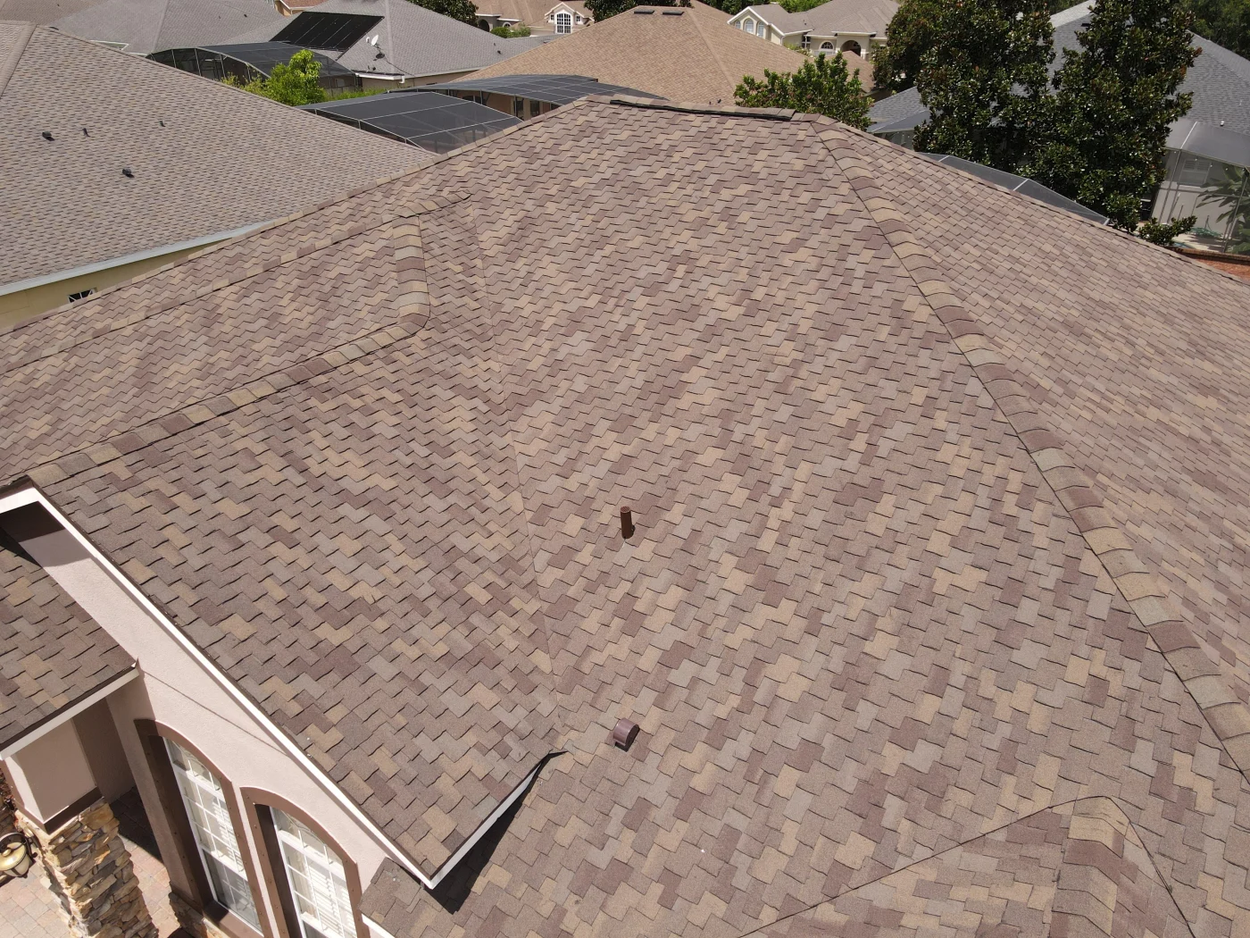 An aerial view of a residential roof covered in brown and tan asphalt shingles, with a visible vent pipe and surrounding houses in the background.