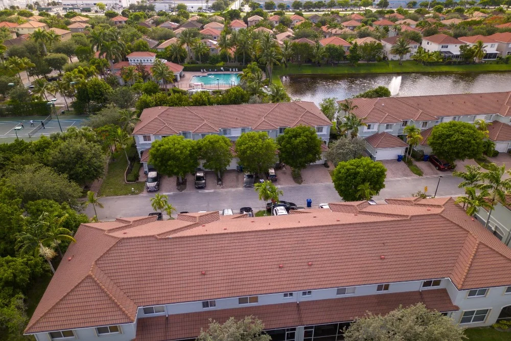 An aerial view of a residential multi-family complex, featuring brown tile roofing, tree-lined streets, tennis courts, and a central swimming pool.