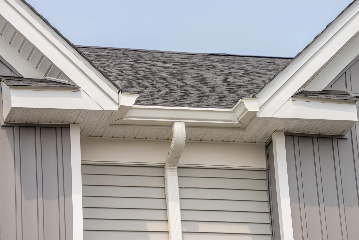 A close-up view of a house's roofline, showing gray shingles, white gutters and downspout, and light gray with vertical and horizontal panels.
