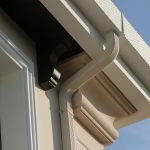 A close-up view of a light-colored gutter system on a building with a textured roof edge and a dark soffit, set against a clear blue sky.