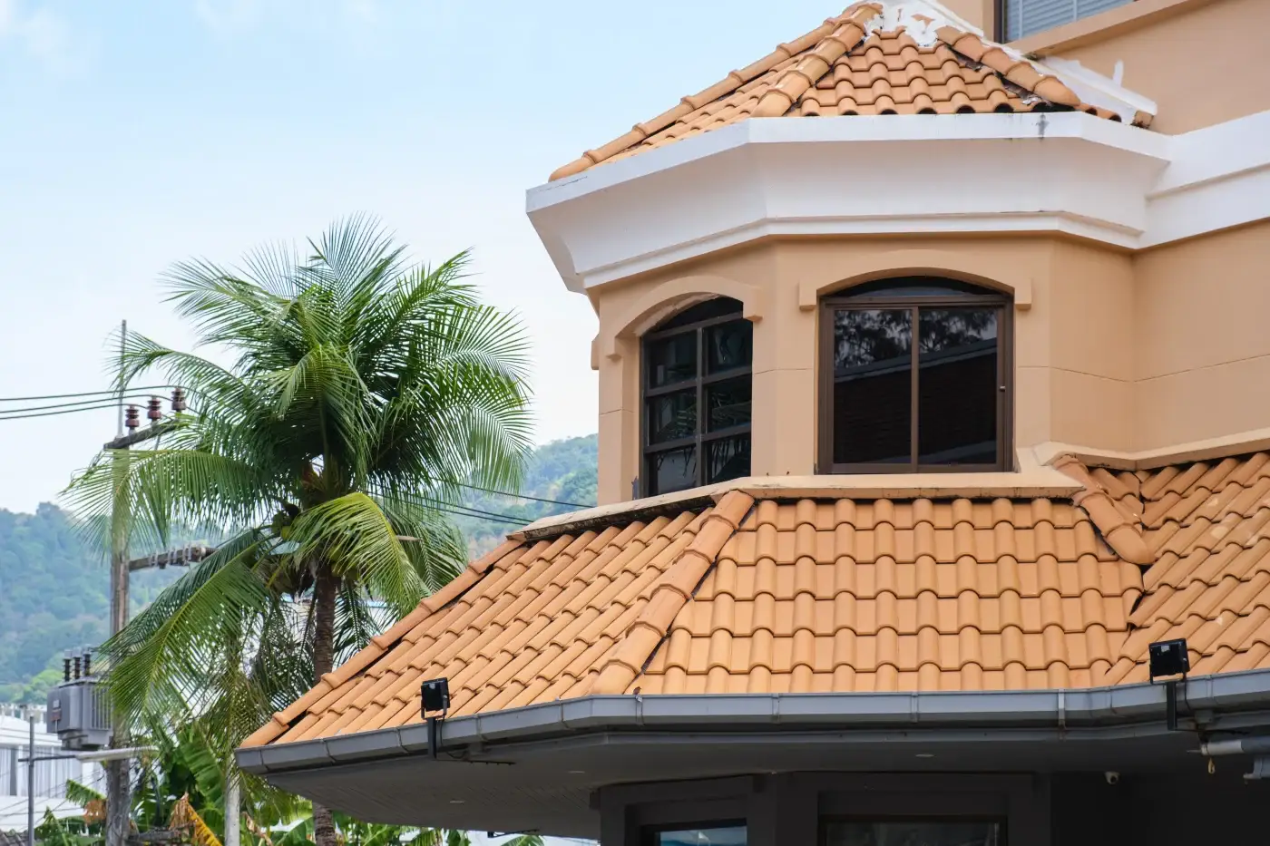 A close-up view of a home with a light beige exterior and a brown-tiled roof, featuring blue hurricane shutters on its windows.