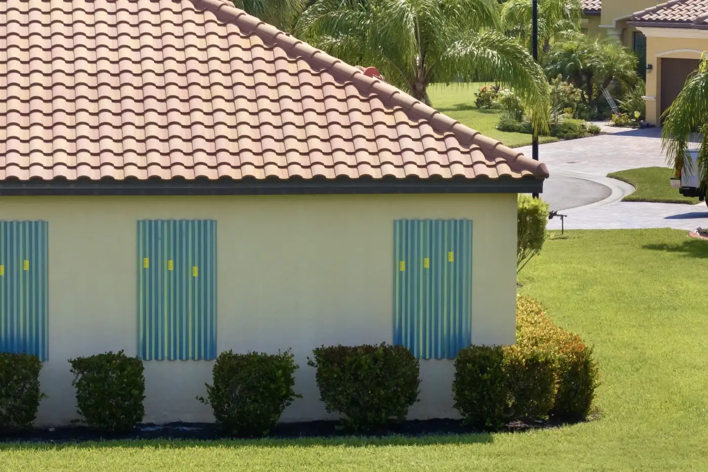 A close-up of a tan-colored building with a light orange clay tile roof, featuring an arched window on the second floor and a palm tree.