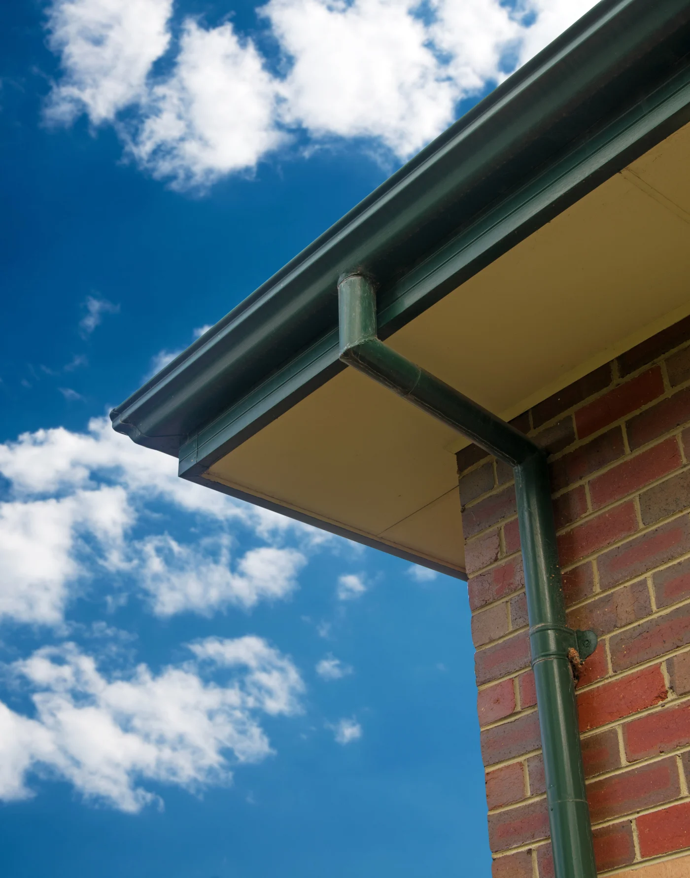 Beautiful Siding, Soffit, Fascia, and Gutters | Fernandina Beach, FL Close-up view of a home’s gutter system attached to a brick exterior wall, showing a green downspout and soffit against a bright blue sky.