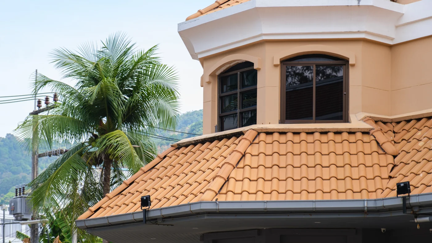 Captivating Tile Roof | Bradenton, FL | Universal Roof and Contracting Close-up view of a tan stucco building with a rounded upper window and a terracotta tile roof, framed by a palm tree and light blue sky in the background.
