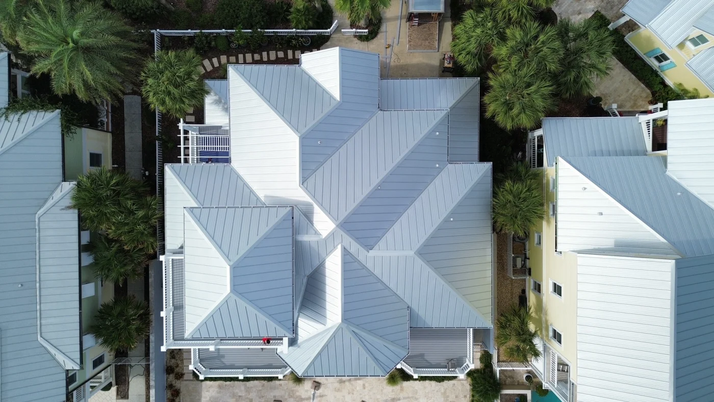 Durbale Metal Roof | Bradenton, FL | Universal Roof and Contracting Aerial view of a coastal-style home featuring an intricate light gray metal roofing system with multiple ridgelines, surrounded by palm trees and neighboring houses.