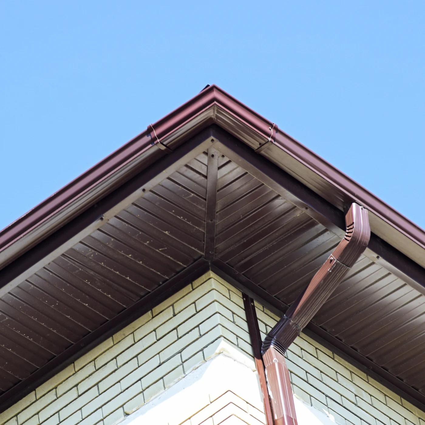 Impeccable Siding Soffit, Fascia, and Gutters | Bradenton, FL Close-up view of a home's brown soffit, fascia, and matching gutter system installed along a brick exterior under a clear blue sky.
