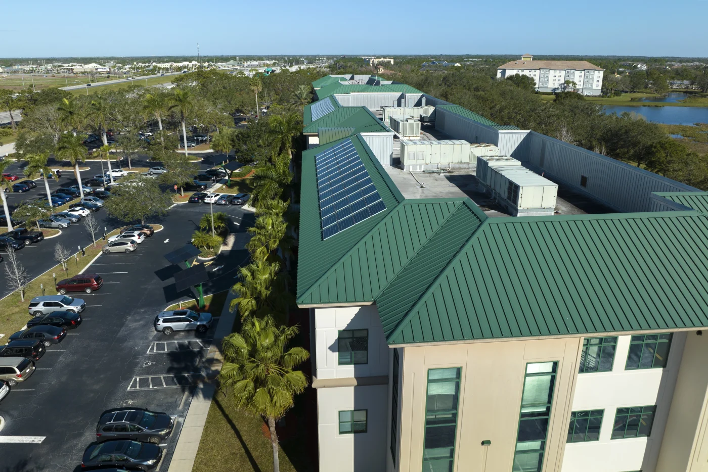 Incredible Commercial Roofing | Bradenton, FL | Universal Roof and Contracting Aerial view of a commercial building with a green metal roof featuring solar panels, surrounded by palm trees, a large parking lot, and nearby residential buildings.