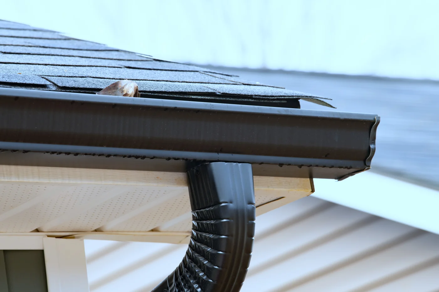 Ravishing Siding, Soffit, Fascia, and Gutters | Riverview, FL | Universal Roof and Contracting Close-up of a home's gutter system showing a dark metal gutter and curved downspout beneath an overhanging roof with damaged asphalt shingles.