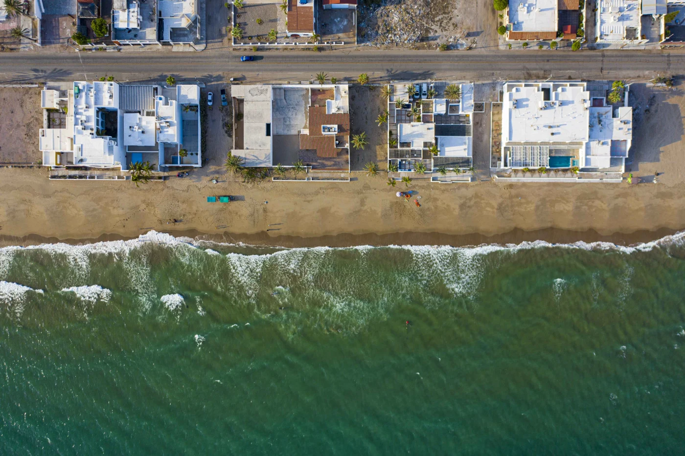 Reliable Commercial Roofing | Bradenton, FL | Universal Roof and Contracting Aerial view of beachfront homes lined along a coastal road, with waves washing onto a sandy shore and clear green ocean water in the foreground.