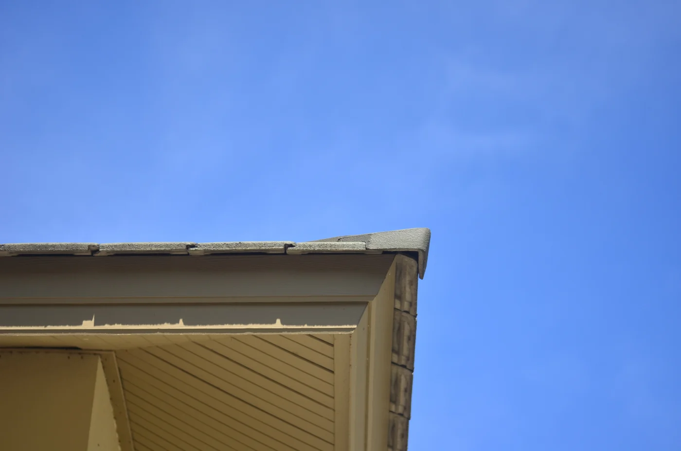 Terrific Siding, Soffit, Fascia, and Gutters | Fernandina Beach, FL Close-up view of a home's roof edge and soffit, showing beige fascia boards, angled soffit panels, and shingle overhang beneath a clear blue sky
