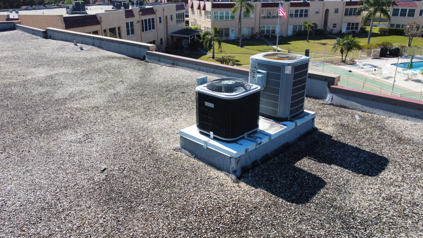 A flat, gravel-covered commercial roof in Central Florida with two large HVAC units installed on a raised platform. In the background, palm trees, a swimming pool, and low-rise residential buildings are visible under a clear blue sky.
