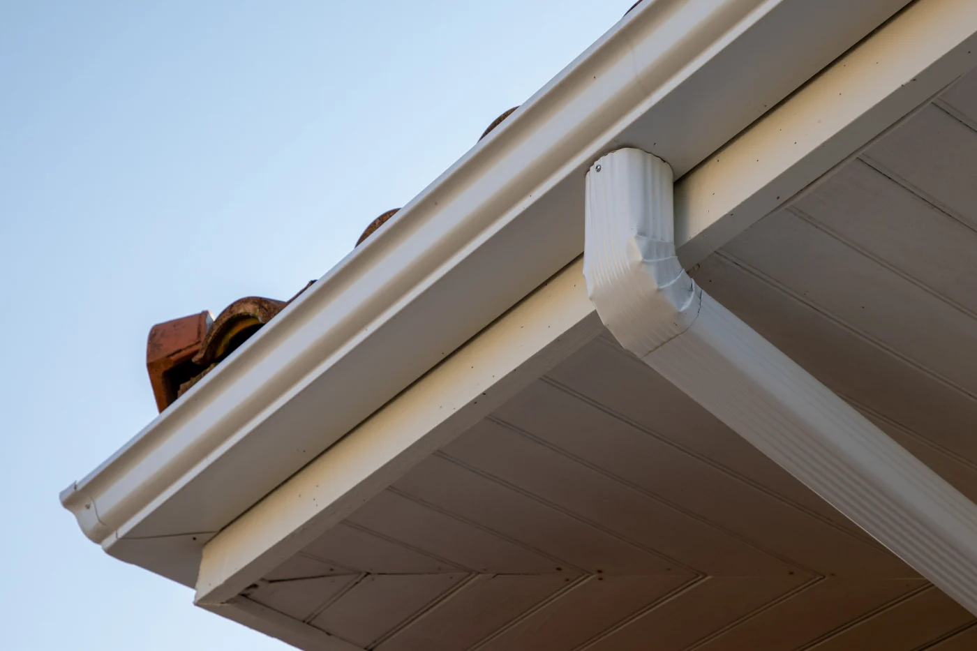 Exceptional Siding, Soffit, Fascia, and Gutters | Windermere, FL | Universal Roof & Contracting Close-up view of a home's white aluminum gutter system and downspout attached beneath the roofline, with terracotta roof tiles and a clean soffit panel