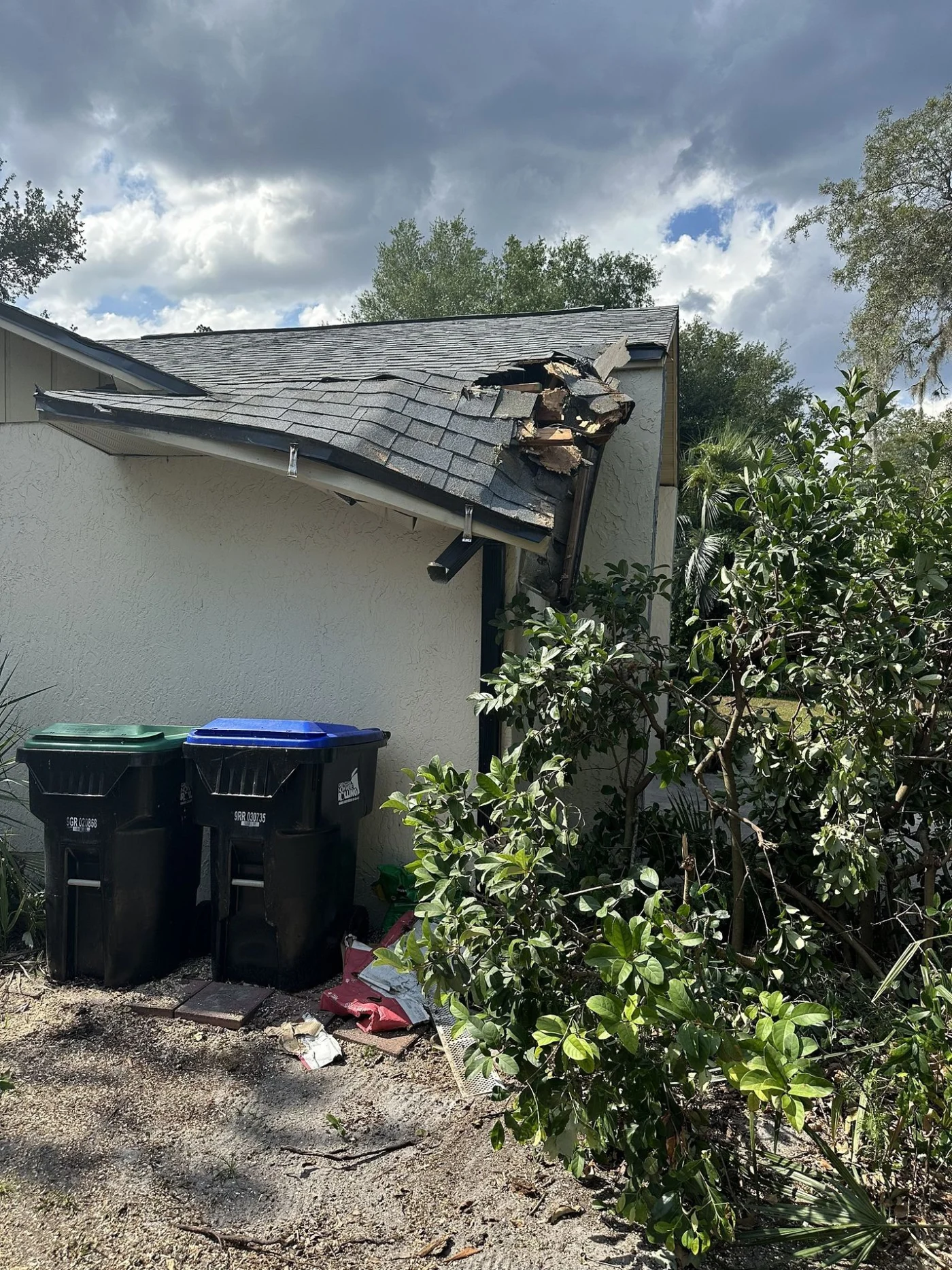 Impeccable Roof Repairs | Winter Park, FL | Universal Roof & Contracting Damaged residential roof with a collapsed corner showing exposed wood and broken shingles, located above two garbage bins.