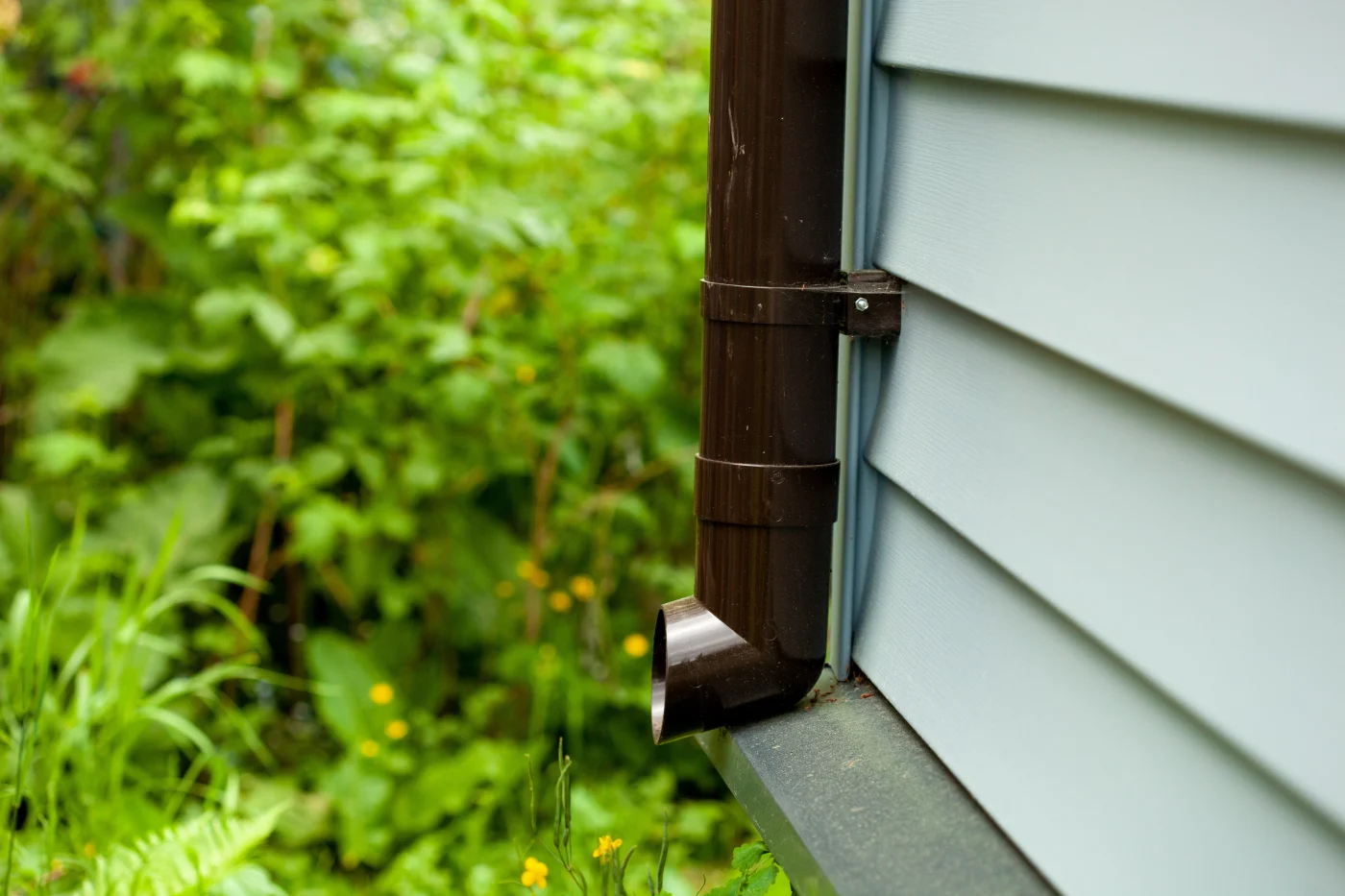 Impressive Siding Soffit Fascia And Gutters Cape Canaveral Fl Universal Roof And Contracting Close-up view of a brown downspout attached to light blue home siding, directing water toward a lush garden area filled with green plants.