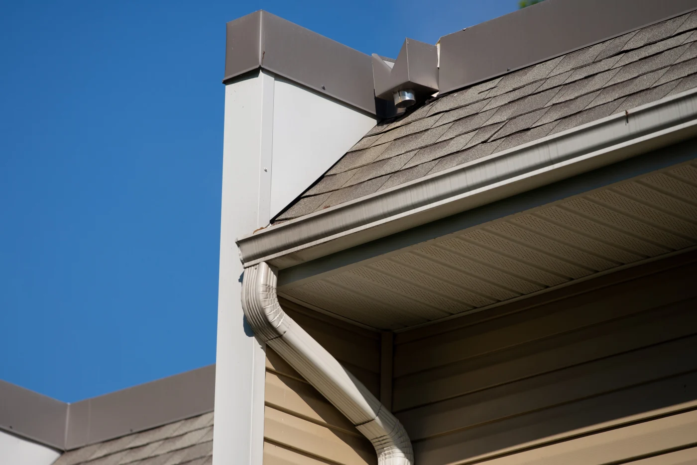 Professional Siding, Soffit, Fascia, and Gutters | Winter Park, FL Close-up view of a home's exterior showing asphalt shingles, beige vinyl siding, perforated soffit panels, and a light-colored downspout