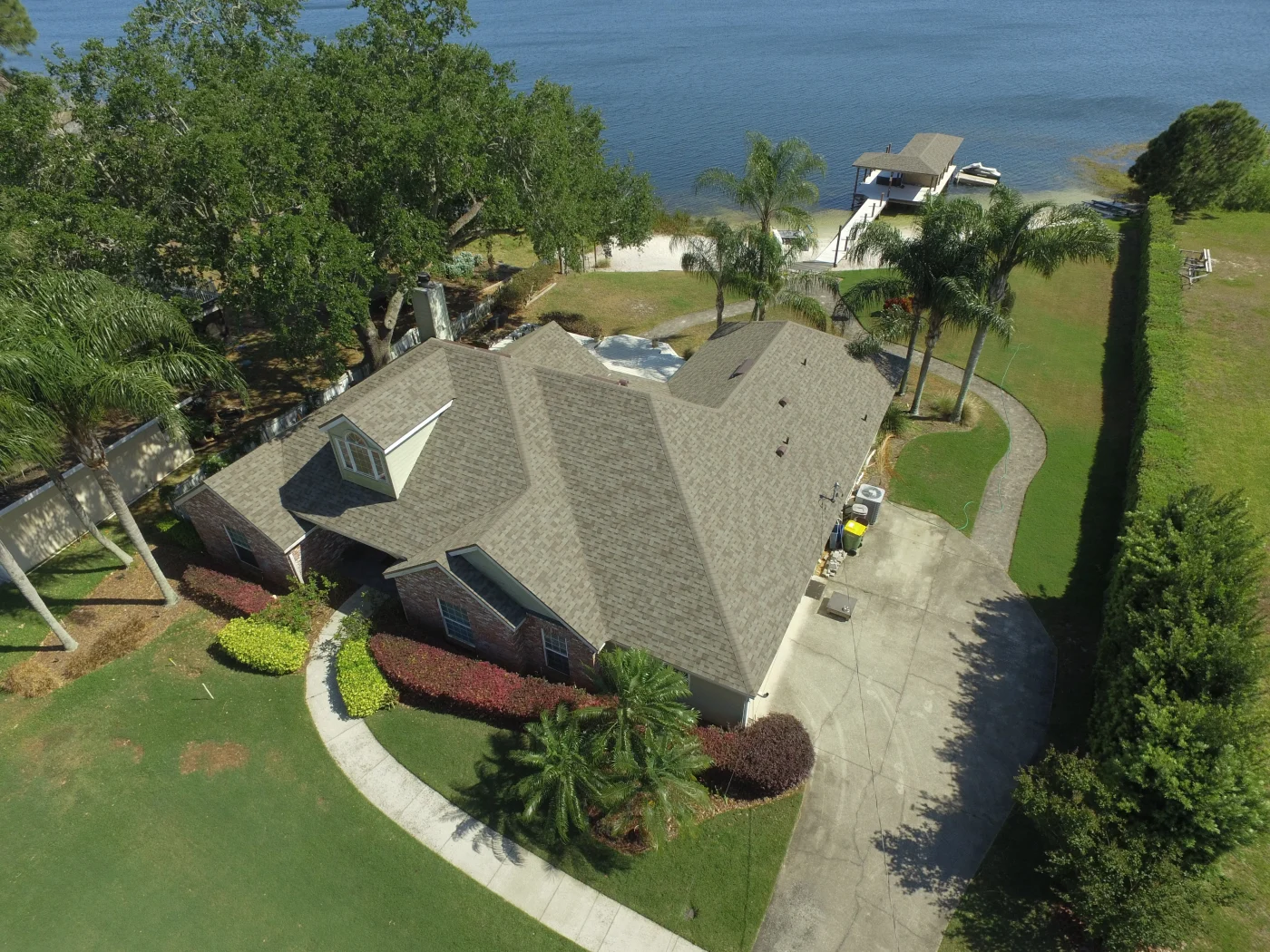 Stellar Asphalt Shingles | Apopka, FL | Universal Roof Aerial view of a lakeside home with a light brown asphalt shingle roof, surrounded by palm trees, landscaped shrubs, and a curved walkway.