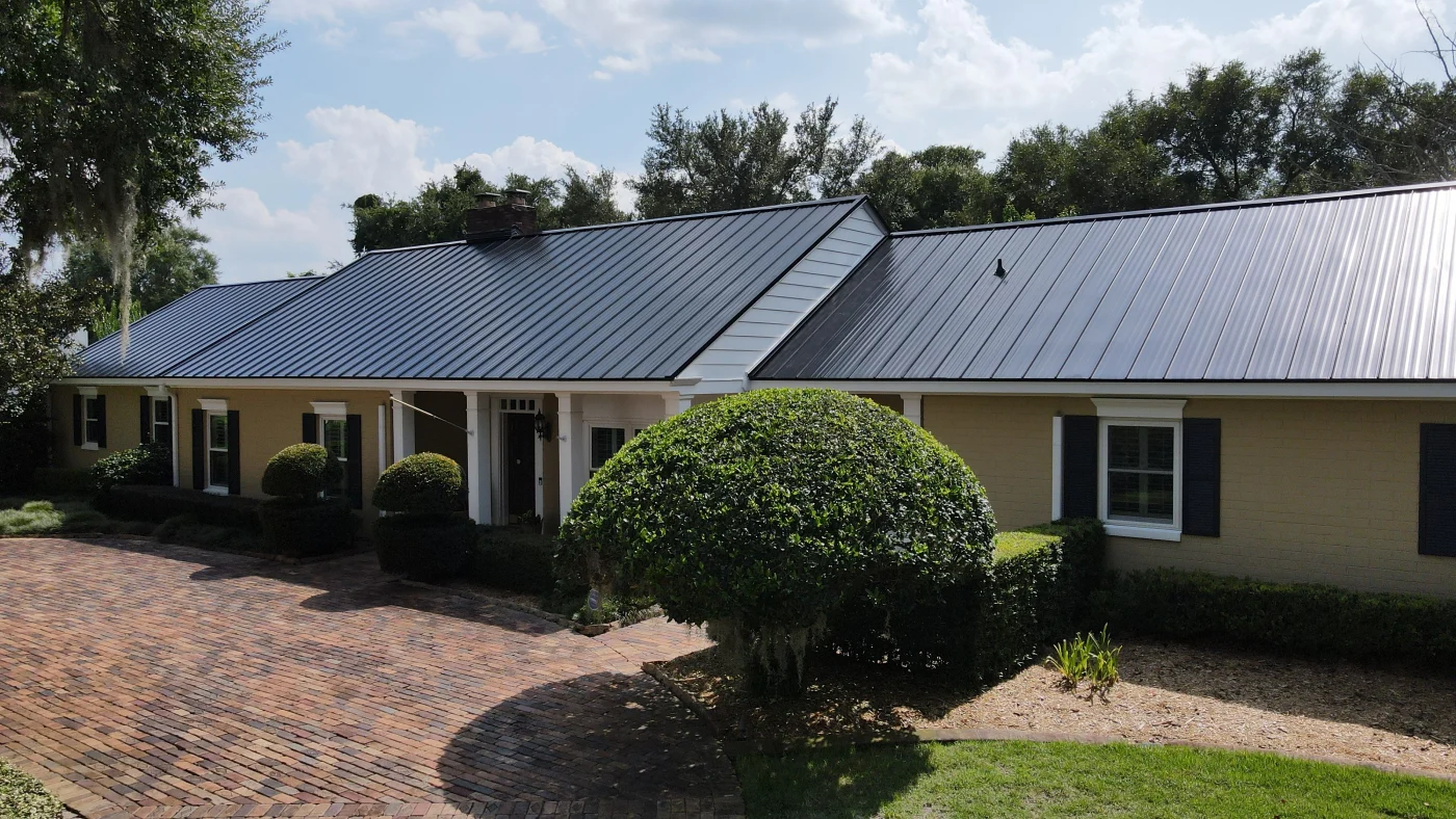 Expert Residential Roofing Services | Ponte Vedra Beach, FL | Universal Roof and Contracting Single-story tan brick home with a sleek black standing seam metal roof, brick paver driveway, and neatly trimmed hedges and shrubs.