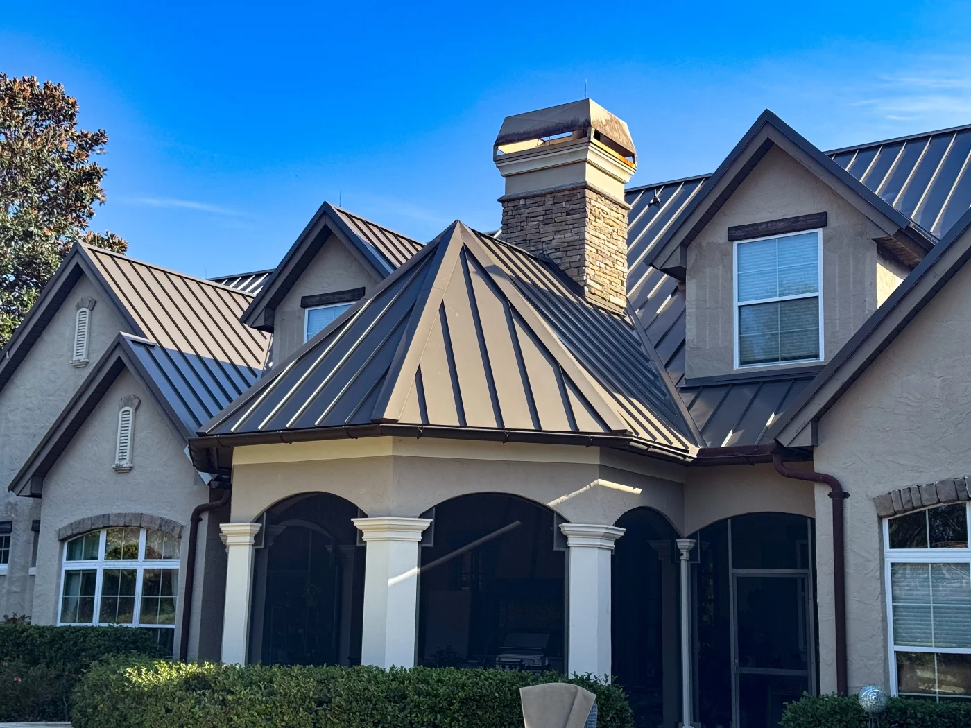 Fabulous Residential Roofing Services | Westchase, FL | Universal Roof and Contracting Front view of an elegant stucco home with a brown standing-seam metal roof, stone chimney, arched covered patio, and white columns.