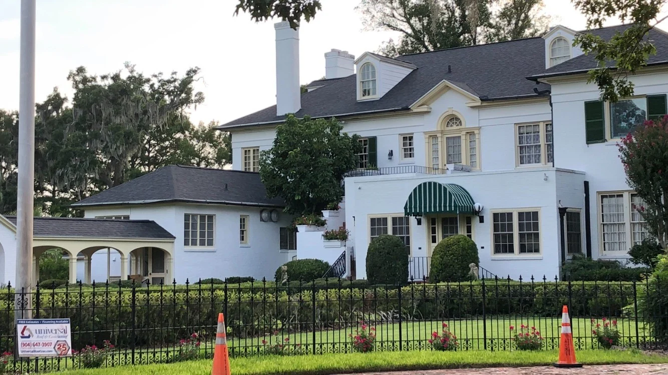 Immaculate Residential Roofing Services | Westchase, FL | Universal Roof and Contracting Elegant historic-style white brick home with a dark shingle roof, green-and-white striped entry awning, and manicured hedges.
